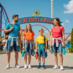 Rodina u vstupu do Energylandie v pohodlném oblečení, vedle barevných horských drah za jasného dne.