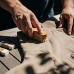 Osoba čistí béžovou semišovou bundu jemným štětcem na dřevěném stole za denního světla.