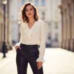 Mladá žena v černých kožených kalhotách a krémové blůze stojí ve městě, pozadí rozmazané.