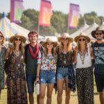 Skupina mladých lidí na festivalu v pohodlném oblečení s doplňky a vodou v ruce, venku za slunečného dne