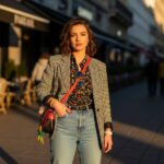 Mladá žena s originálním oblečením stojí ve městě za zlaté hodiny, okolí kaváren a lidí rozmazané.