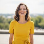 Šťastná žena v žlutých šatech venku na slunci s městským pozadím a jemným větrem ve vlasech