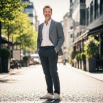 Muž v elegantním smart casual oblečení stojí ulice s kavárnami, s rukama v kapsách a lehkým úsměvem.